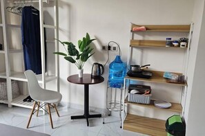 Een kleine kamer met een witte stoel, een klein rond tafeltje met een plant, een waterdispenser en planken met diverse spullen.