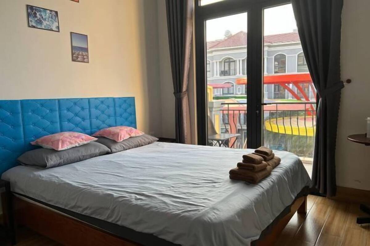 A bedroom with a blue headboard, a bed with white sheets, and a view of buildings through a glass door.