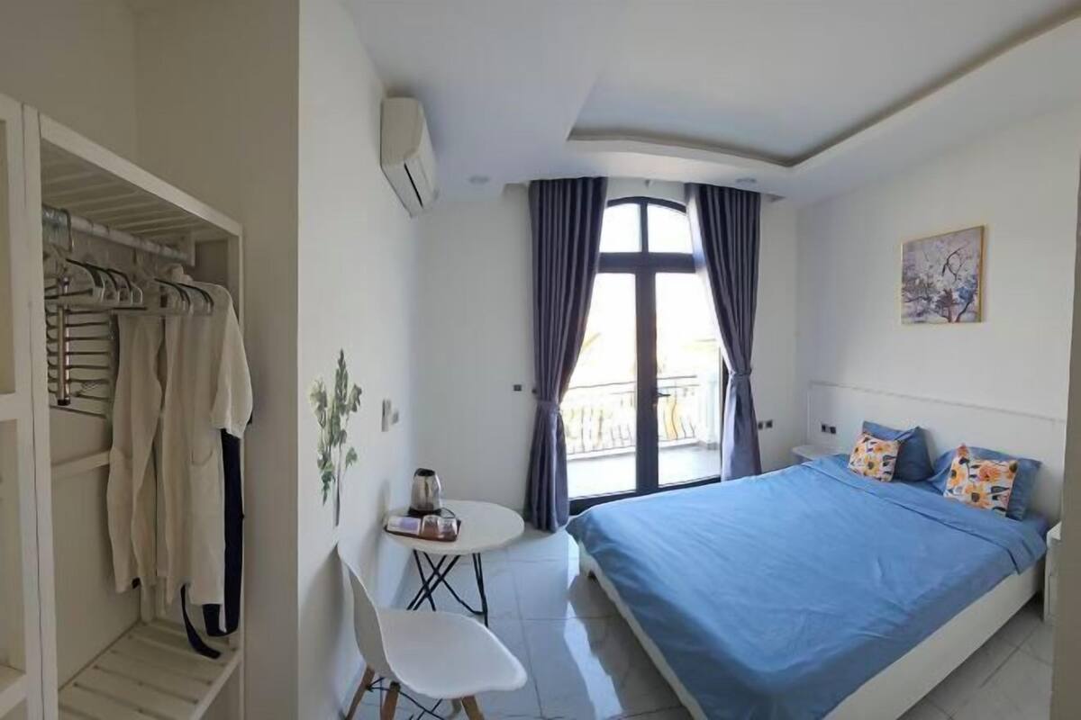 A bedroom with a bed, a chair, a small table, a window with curtains, and a wall-mounted air conditioner.