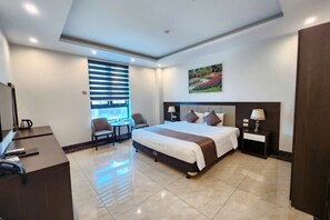 A hotel room with a large bed, bedside lamps, a desk with a chair, a window with blinds, and a painting on the wall. - LAVANDER HOTEL (Bac Ninh)