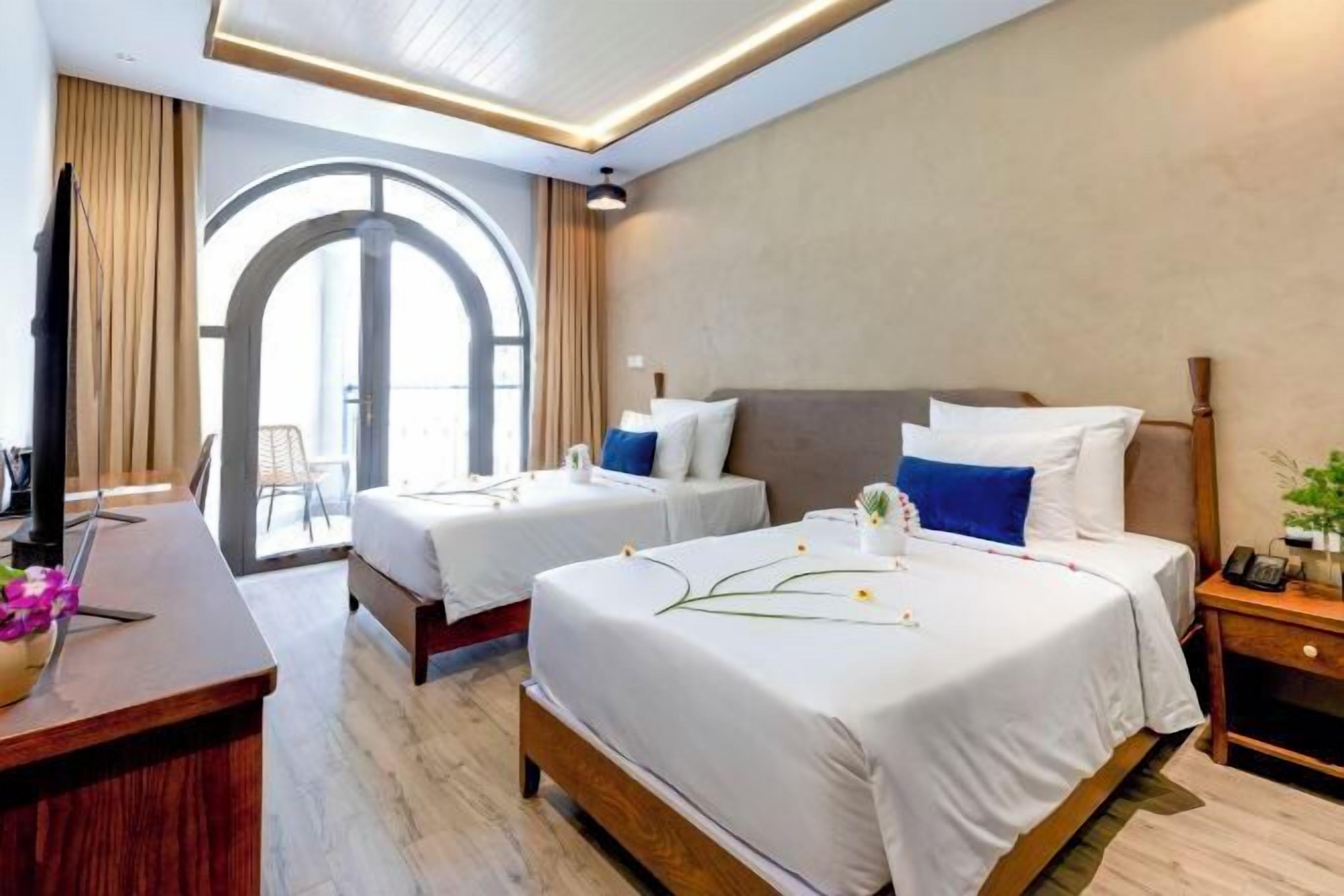 A hotel room with two beds, a desk, a chair, a TV, and a large arched window.
