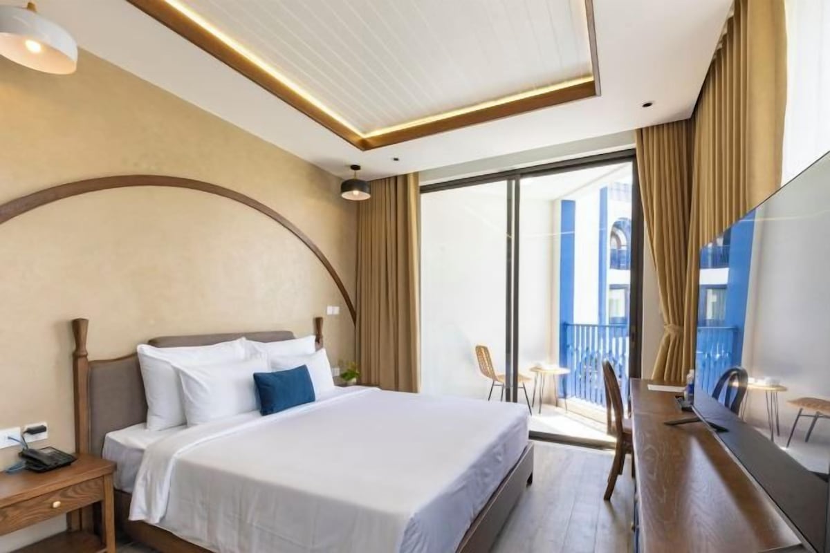 A modern hotel room with a large bed, a desk, a flat-screen TV, and a balcony with blue railings.
