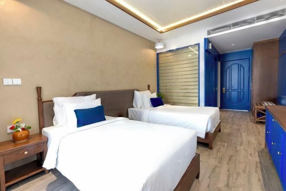 A hotel room with two beds, a wooden floor, a blue door, and a small table with a vase.
