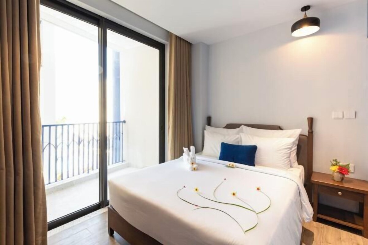 A hotel room with a large bed, a nightstand with a vase of flowers, and a balcony with a railing.