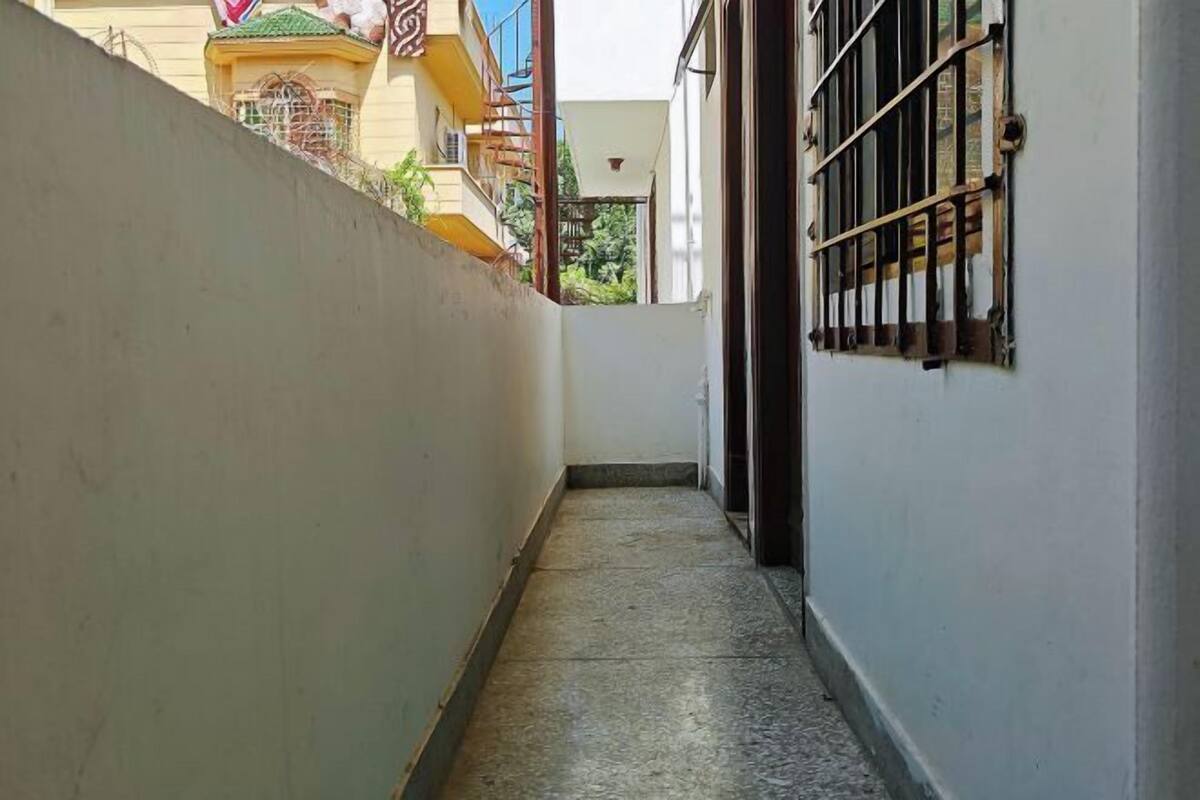 A narrow corridor with a tiled floor, a white wall on one side, and a building with barred windows on the other.