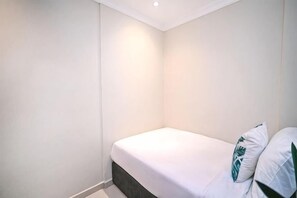 Una habitación pequeña con cama individual, cubrecama y almohadas blancas, cabecera oscura y luz empotrada en el techo.