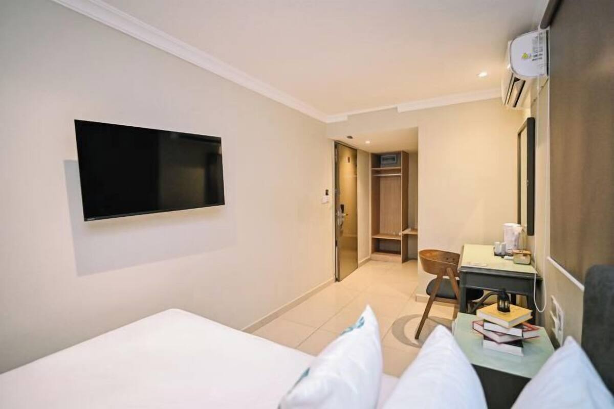 A hotel room with a bed, a desk with a chair, a television mounted on the wall, and a wardrobe.