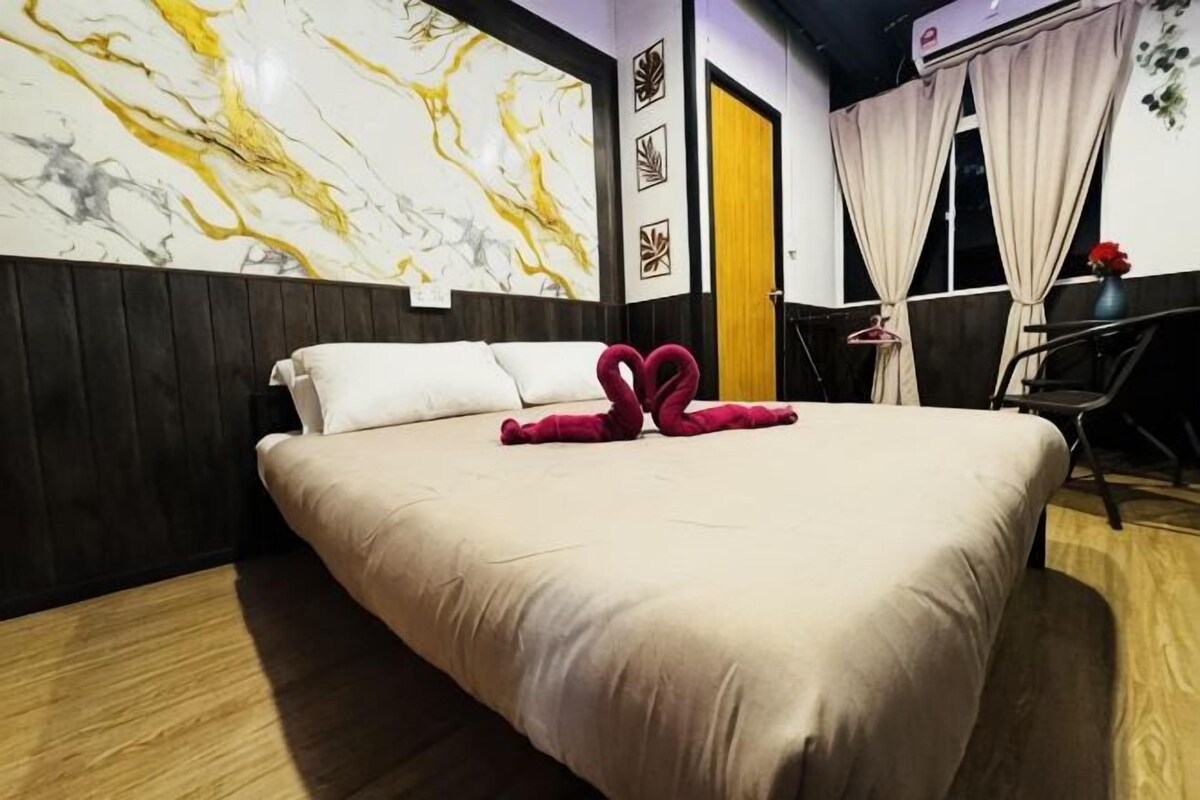 A hotel room with a bed, a wooden headboard, a marble wall, a window with curtains, and a small table with a vase.