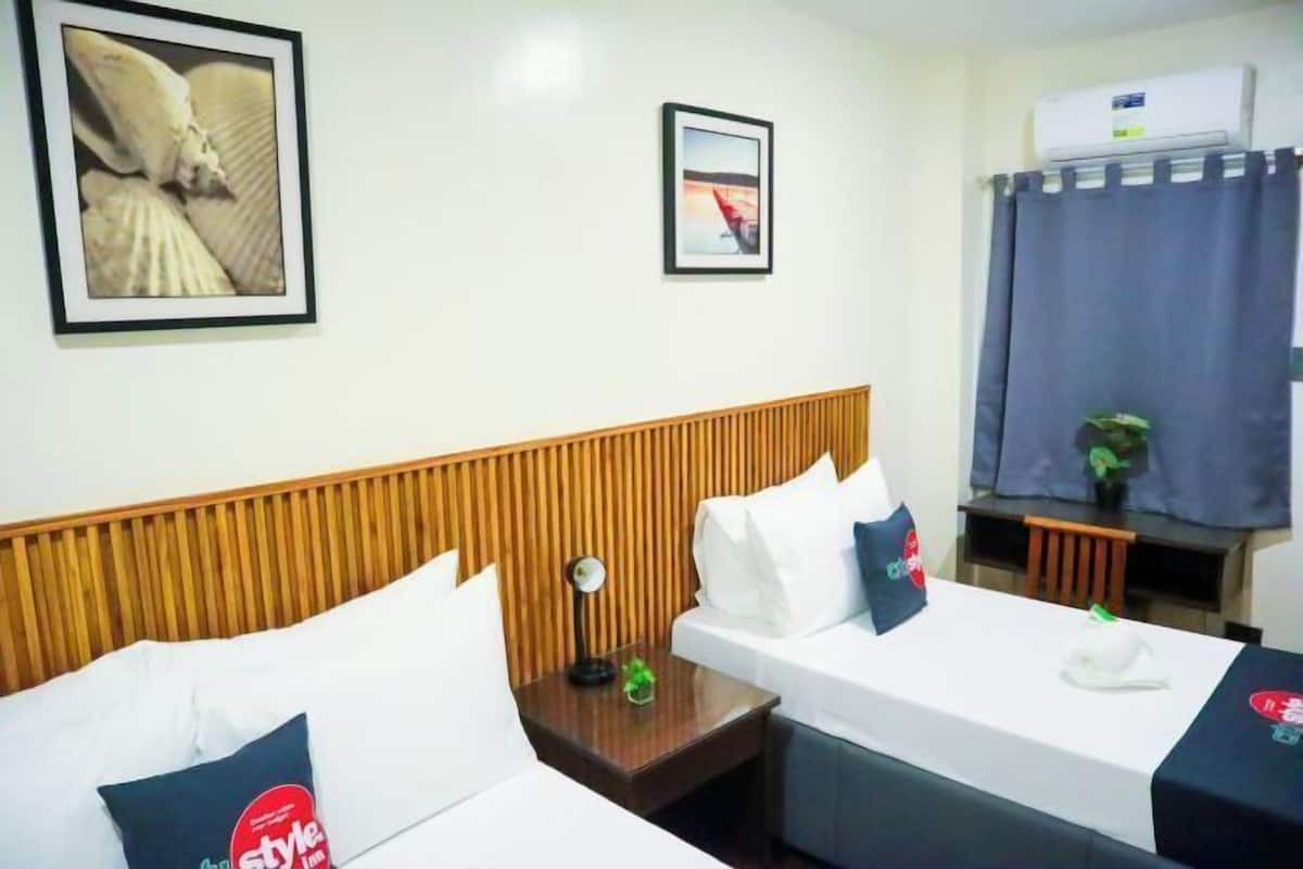 A hotel room with two beds, a wooden headboard, a small table with a lamp, a framed picture on the wall, and an air conditioning unit.