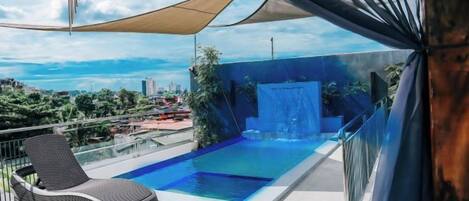 A rooftop infinity pool with a waterfall feature, a lounge chair, and a view of the cityscape.