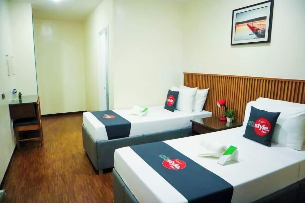 A hotel room with two beds, a wooden headboard, a small table with a plant, and a framed picture on the wall.