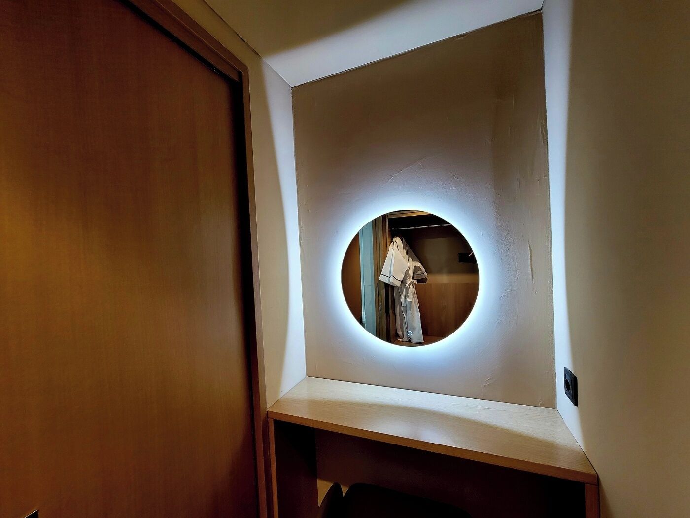Un espejo pequeño y redondo con luz alrededor, fijado a la pared en un rincón de la habitación.
