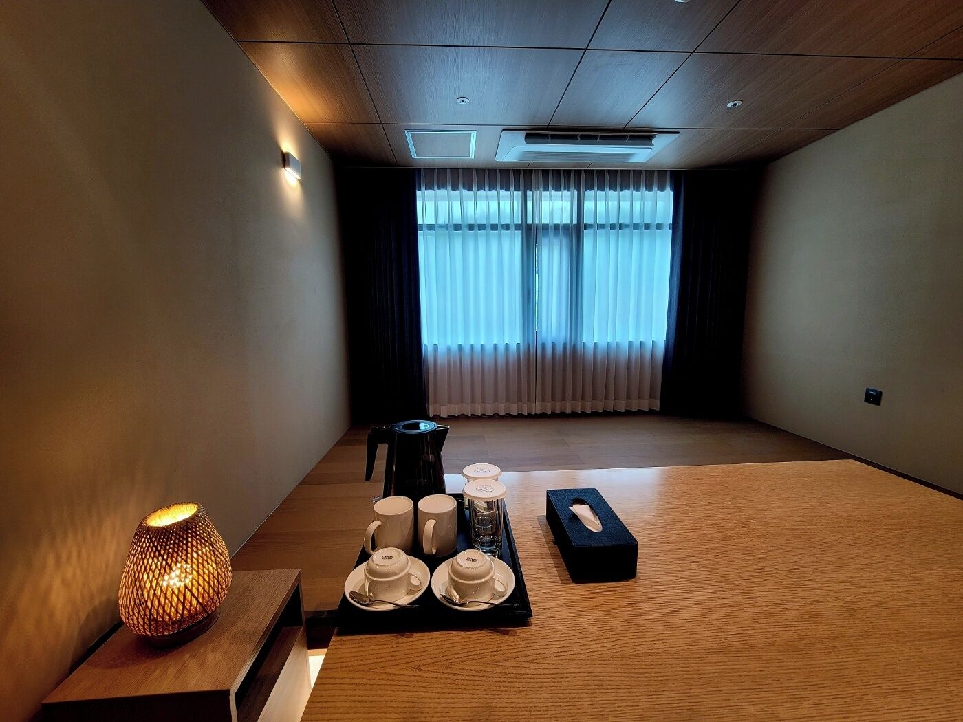A room with a wooden floor, a small table with a tea set, a decorative lamp, and a window with sheer curtains.