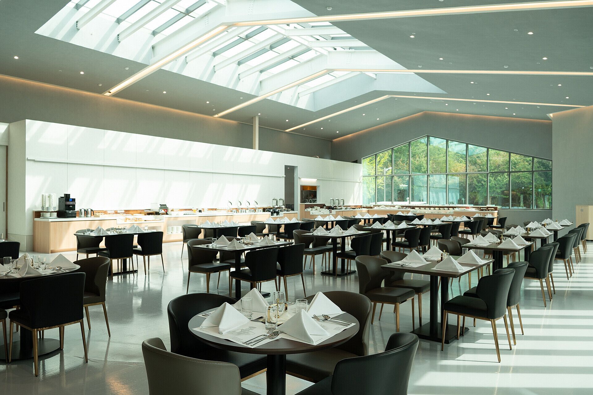 Un espace de restauration moderne avec des tables dressées, une cuisine en arrière-plan et de grandes fenêtres laissant entrer la lumière naturelle.