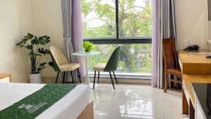 Une chambre d’hôtel avec un lit, une petite table ronde et une chaise, une plante en pot et une vue sur la verdure à l’extérieur.