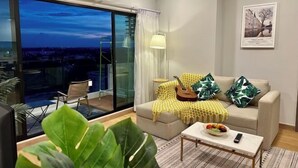 Un salon moderne avec un canapé, une table basse, une guitare et une vue sur la ville au crépuscule.