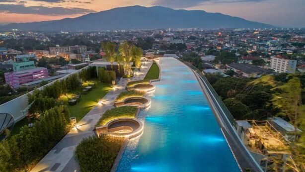 Une piscine au design incurvé, entourée de verdure et de sentiers, avec une vue sur la ville et les montagnes en arrière-plan.