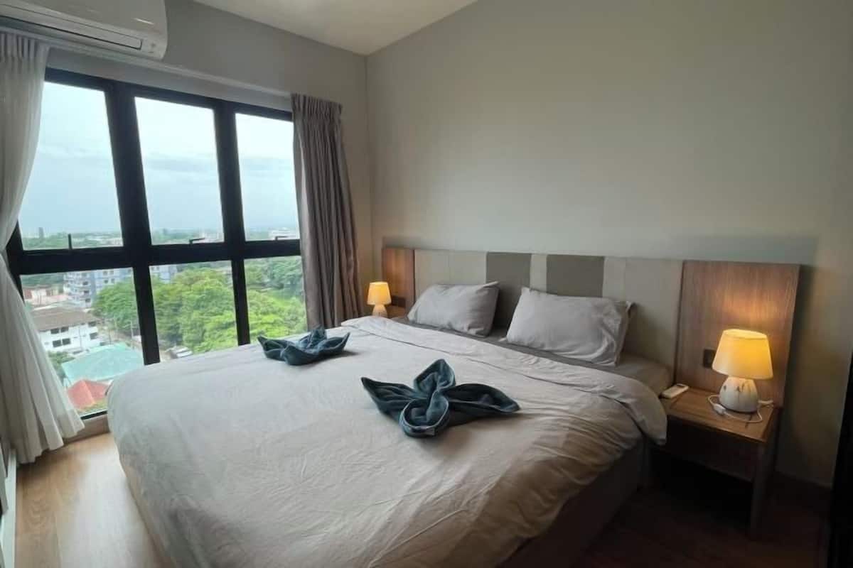 A hotel room with a large bed, two bedside lamps, a window with curtains, and a view of buildings and trees.