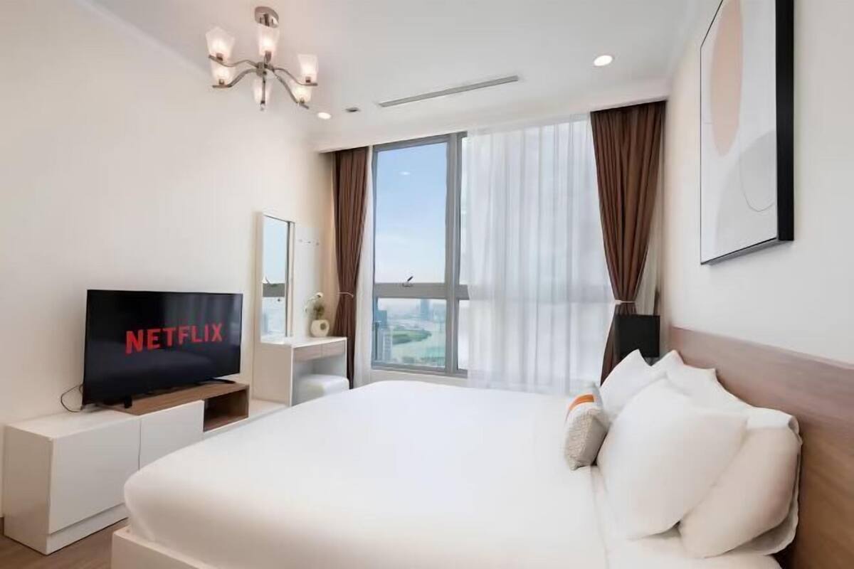 Habitació d'hotel moderna amb un llit gran, una televisió sobre un suport, una taula de nit i una finestra amb cortines.