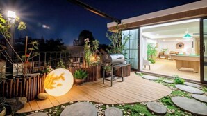 A modern outdoor patio with a grill, wooden deck, and a view into a well-lit interior space. - Alden Design Pool Villa (Pattaya)