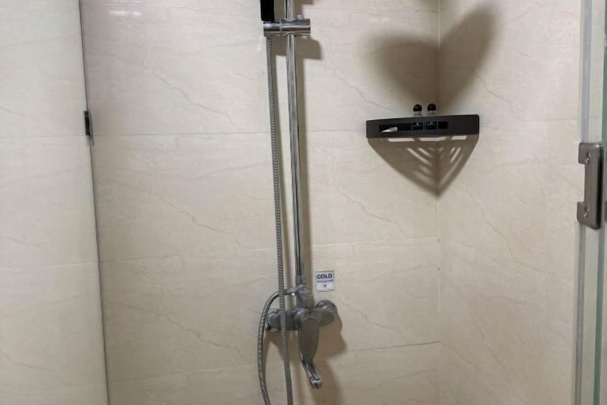 A shower with a handheld showerhead, a wall-mounted control panel, and a small shelf.