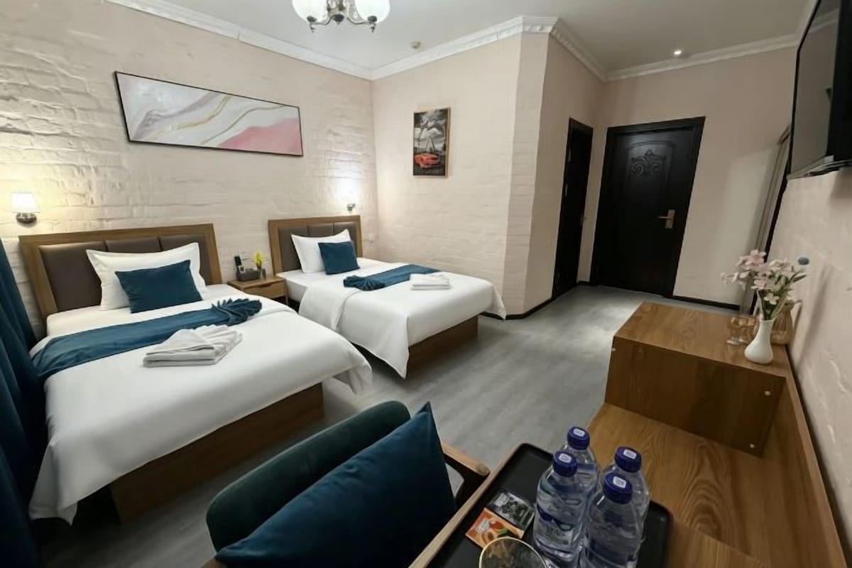 A hotel room with two beds, a desk, and a chair. There is a tray with bottled water and a vase with flowers on the desk.