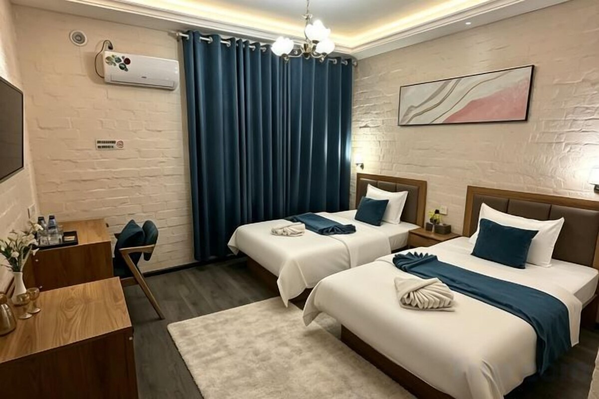 A hotel room with two beds, a desk, a chair, a small table, and a wall-mounted air conditioner.