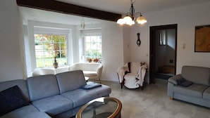 A living room with two sofas, a glass coffee table, a white armchair, and a dining table. There is a large window with a view of greenery outside. - Activity country house near the Baltic Sea with pool & orchard, for family & friends (Lübow)