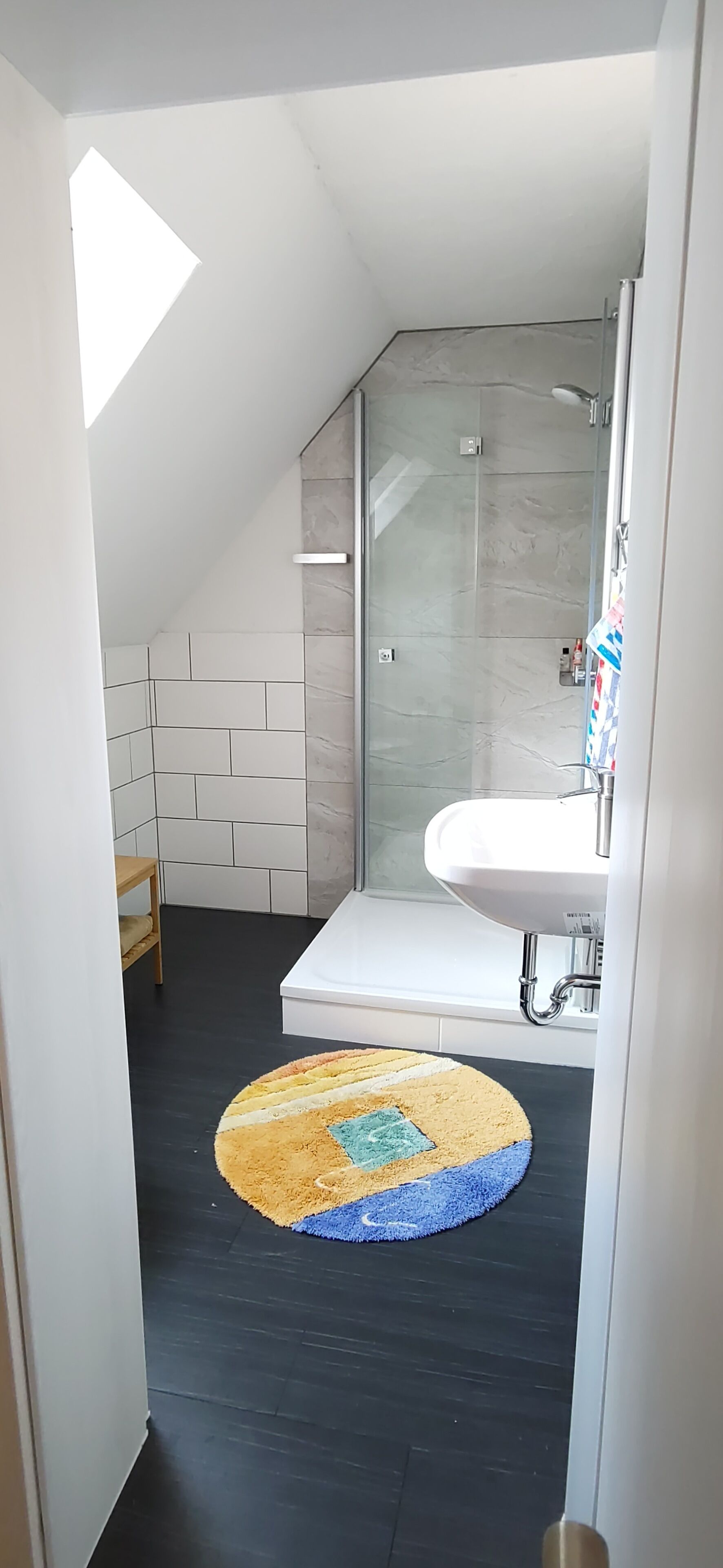 A modern bathroom with a glass shower, white tiles, a white sink, and a colorful rug.