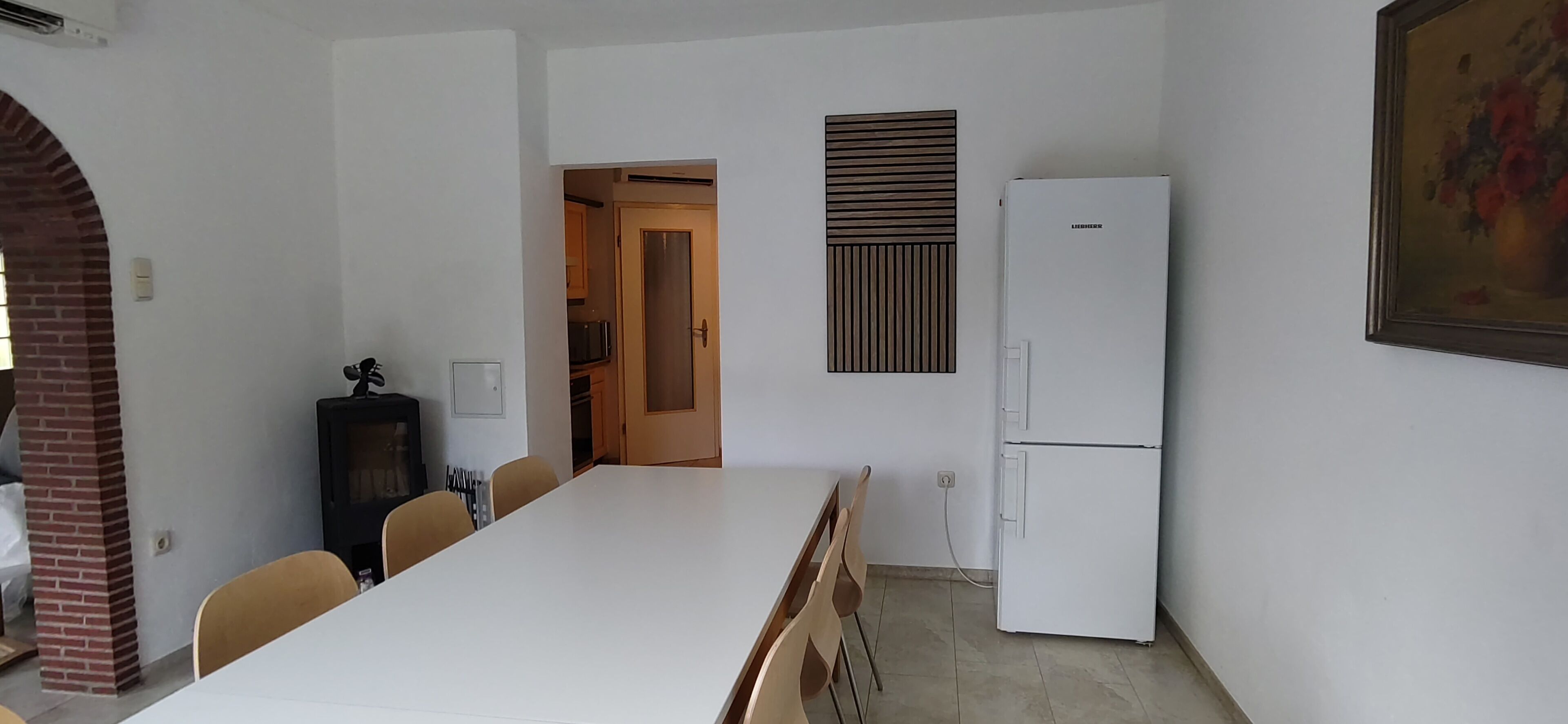 Uma área de refeições com uma mesa comprida, cadeiras, uma geladeira e uma porta que dá para uma cozinha.