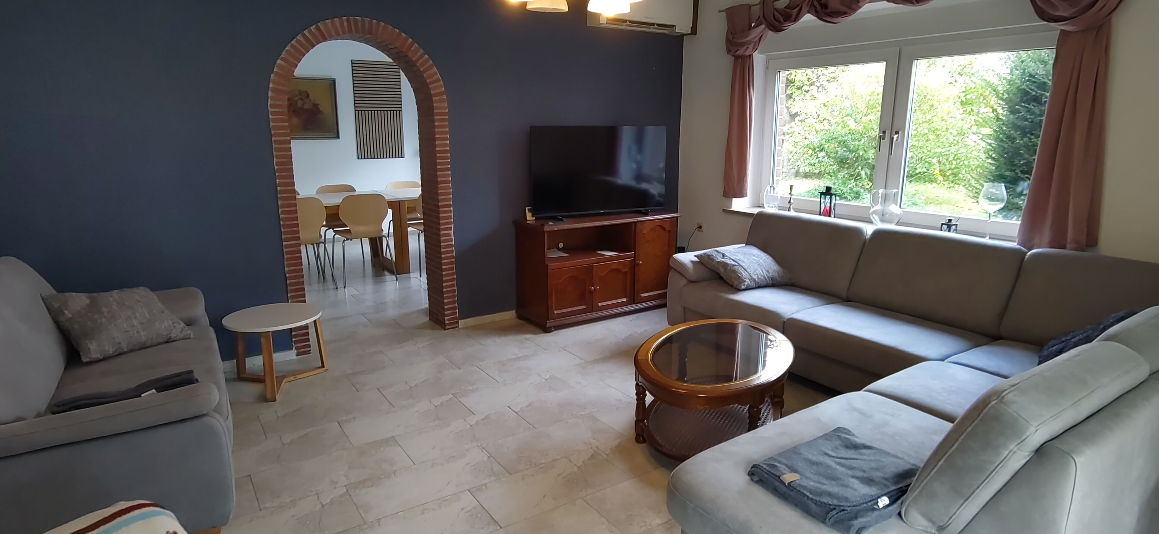 Una sala de estar con sofá, televisión, mesa de comedor con sillas y ventana con cortinas.