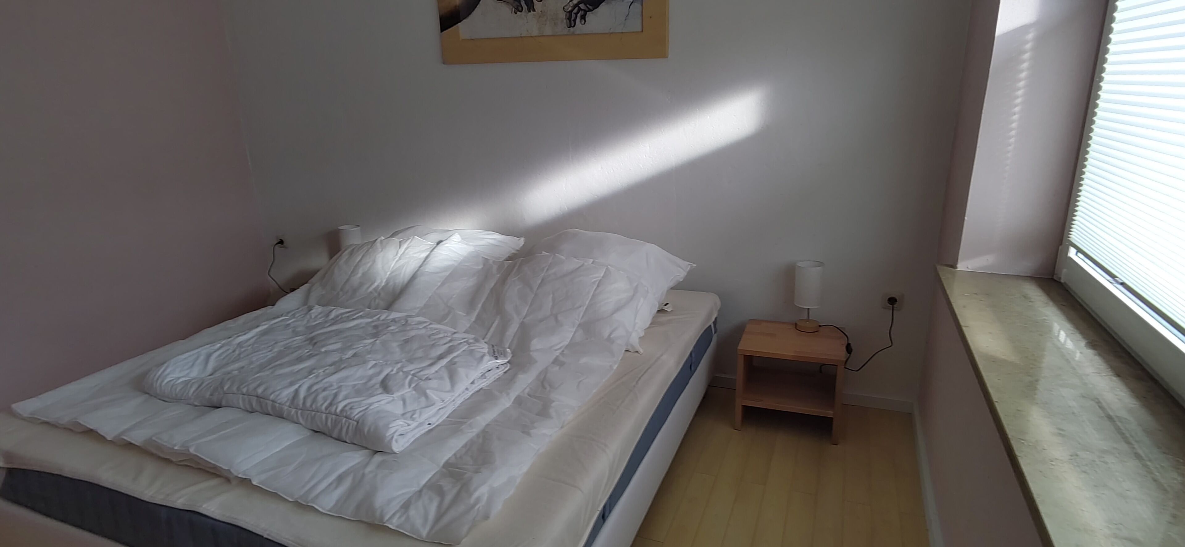 Una cama bien hecha con ropa blanca, una mesita de noche con lámpara y una ventana con persianas.