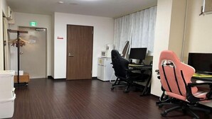A hotel room with a wooden floor, a desk with a computer, two office chairs, a wardrobe, and a door with an exit sign. - Tokyo guest (Tokyo)