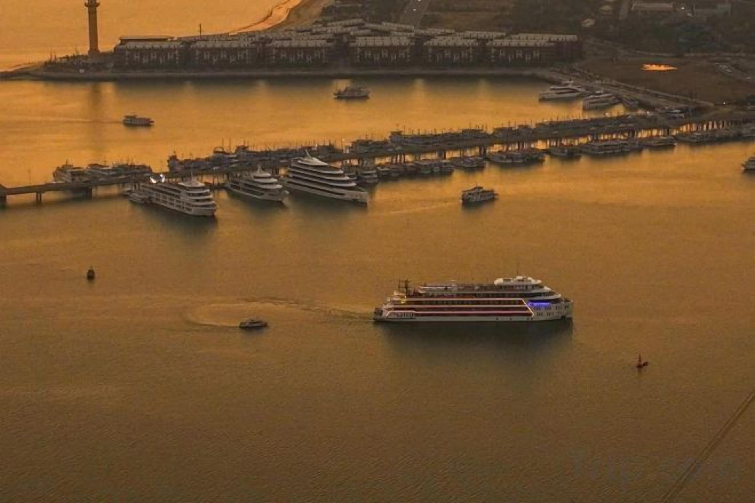 A large cruise ship docked at a port with multiple smaller boats and a lighthouse in the background.