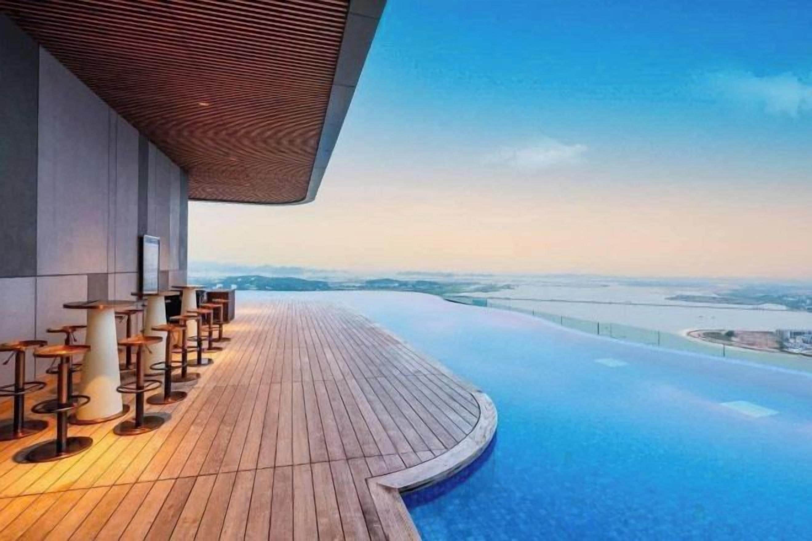 Une piscine à débordement située sur le toit, avec une terrasse en bois et un espace salon, offrant une vue panoramique sur un paysage lointain.