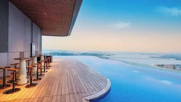 Une piscine à débordement située sur le toit, avec une terrasse en bois et un espace salon, offrant une vue panoramique sur un paysage lointain.