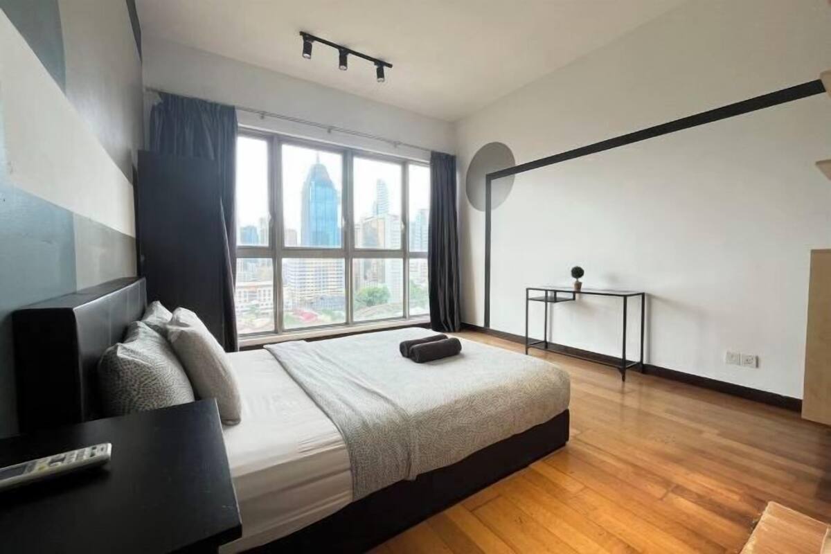 Une chambre moderne avec un grand lit, une petite table, une fenêtre donnant sur la ville et un cadre photo noir accroché au mur.
