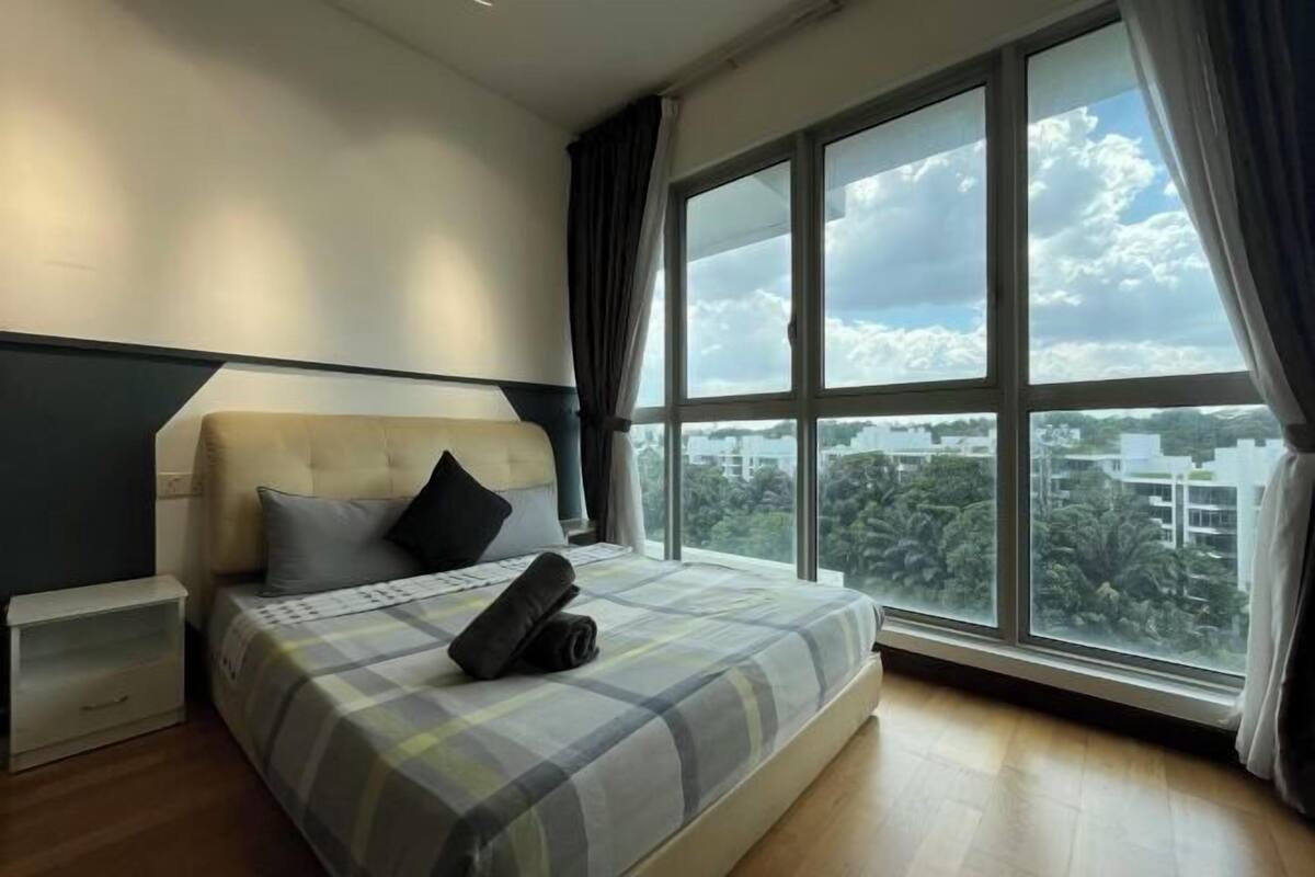 Une chambre avec une grande fenêtre, un lit recouvert d’une couverture à carreaux, une table de chevet et une tête de lit.
