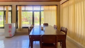 A dining area with a long wooden table and chairs, a refrigerator, and large windows with curtains. - Raja Asri Guest House Sanur Mitra RedDoorz (Bali)