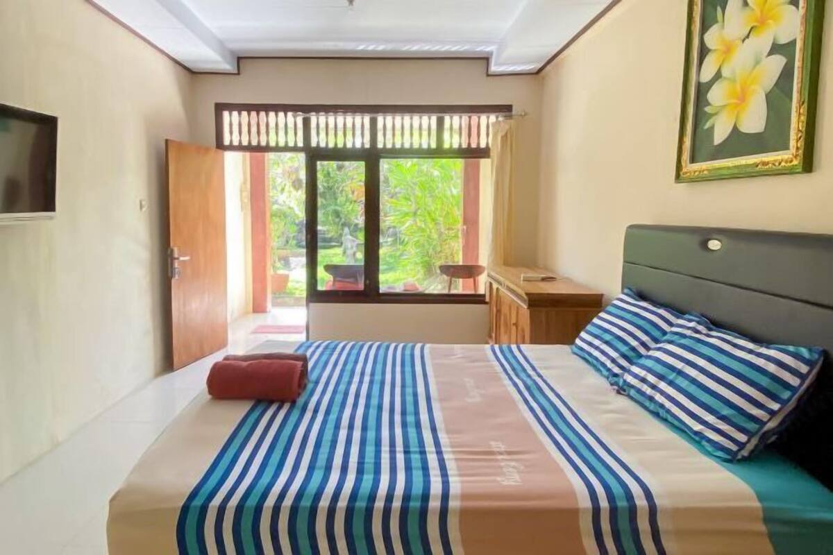 Een slaapkamer met een bed, een nachtkastje, een raam met uitzicht op groen, en een deur.