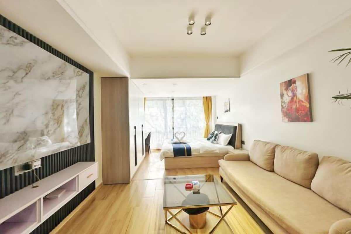 A modern living room with a large TV, a sofa, a glass coffee table, and a bed in the background.