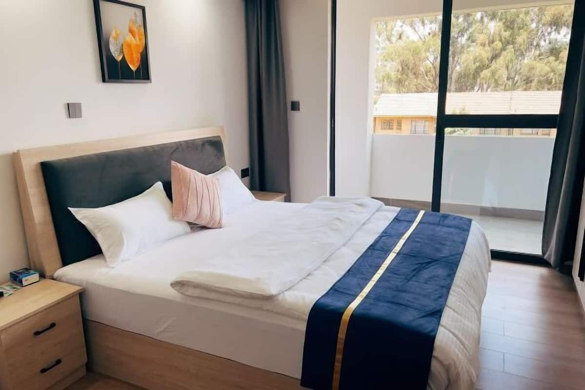 A hotel room with a bed, a nightstand, a window with a view of trees, and a framed picture on the wall.