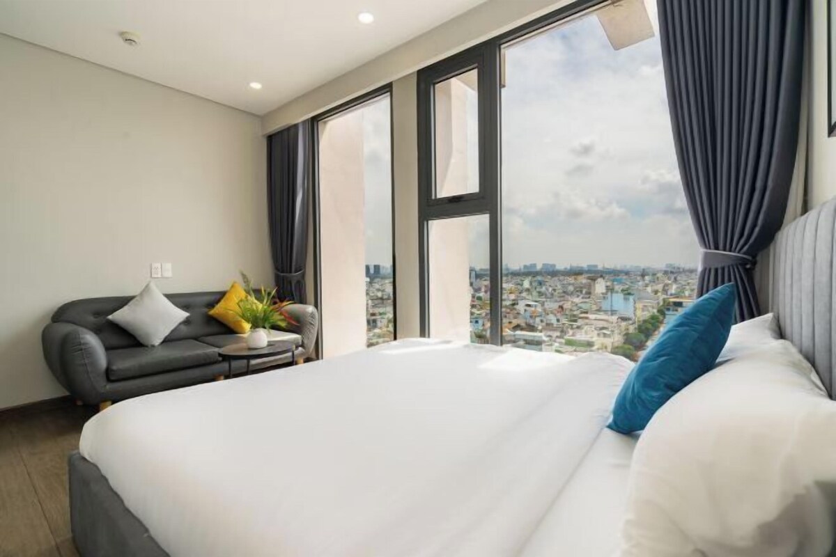 A hotel room with a large bed, a sofa, a small table with a plant, and a city view through large windows.