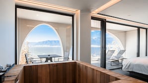 A modern room with a wooden bench, a bed, and large windows offering a view of mountains and a body of water.