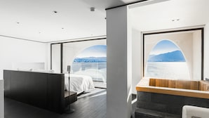 A modern hotel room with a large bed, a desk, and a view of a body of water and cityscape through arched windows.