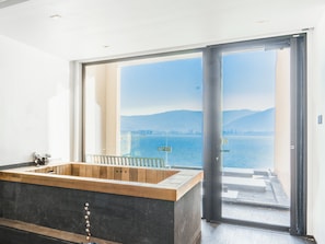 A modern bathroom with a wooden bathtub, a large window, and a view of the sea and mountains.