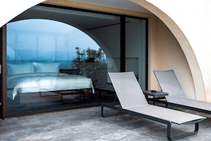 A modern bedroom with a glass door leading to a balcony, a bed, and two lounge chairs.