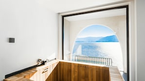 A modern bathroom with a wooden bench, a built-in sink, and a large arched window offering a view of a body of water and distant mountains.