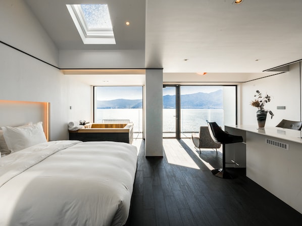 A modern hotel room with a large bed, a sitting area with a view of the water, and a balcony with a cityscape view.