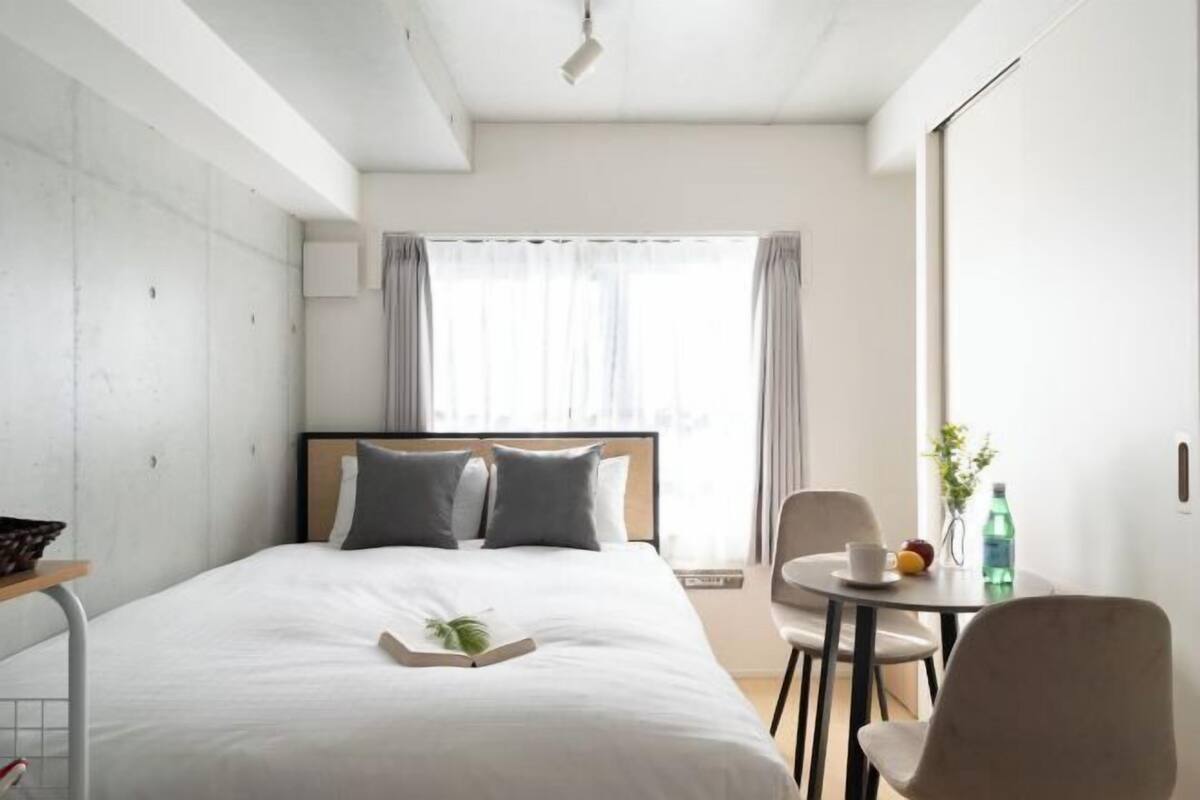 A modern hotel room with a large bed, a bedside table, a small round table with a chair, and a window with curtains.